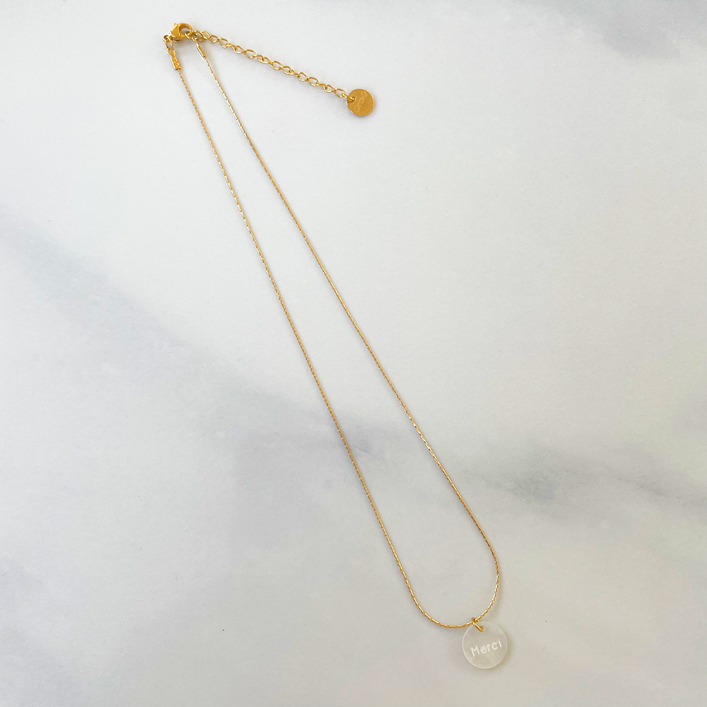Une photo mettant en valeur un collier élégant en acier inoxydable, orné d'un pendentif délicat en nacre affichant le mot "merci" avec finesse. L'acier contraste harmonieusement avec la douceur de la nacre, créant ainsi un bijou raffiné et expressif. Ce collier incarne à la fois la grâce et la gratitude dans un design minimaliste et chic.