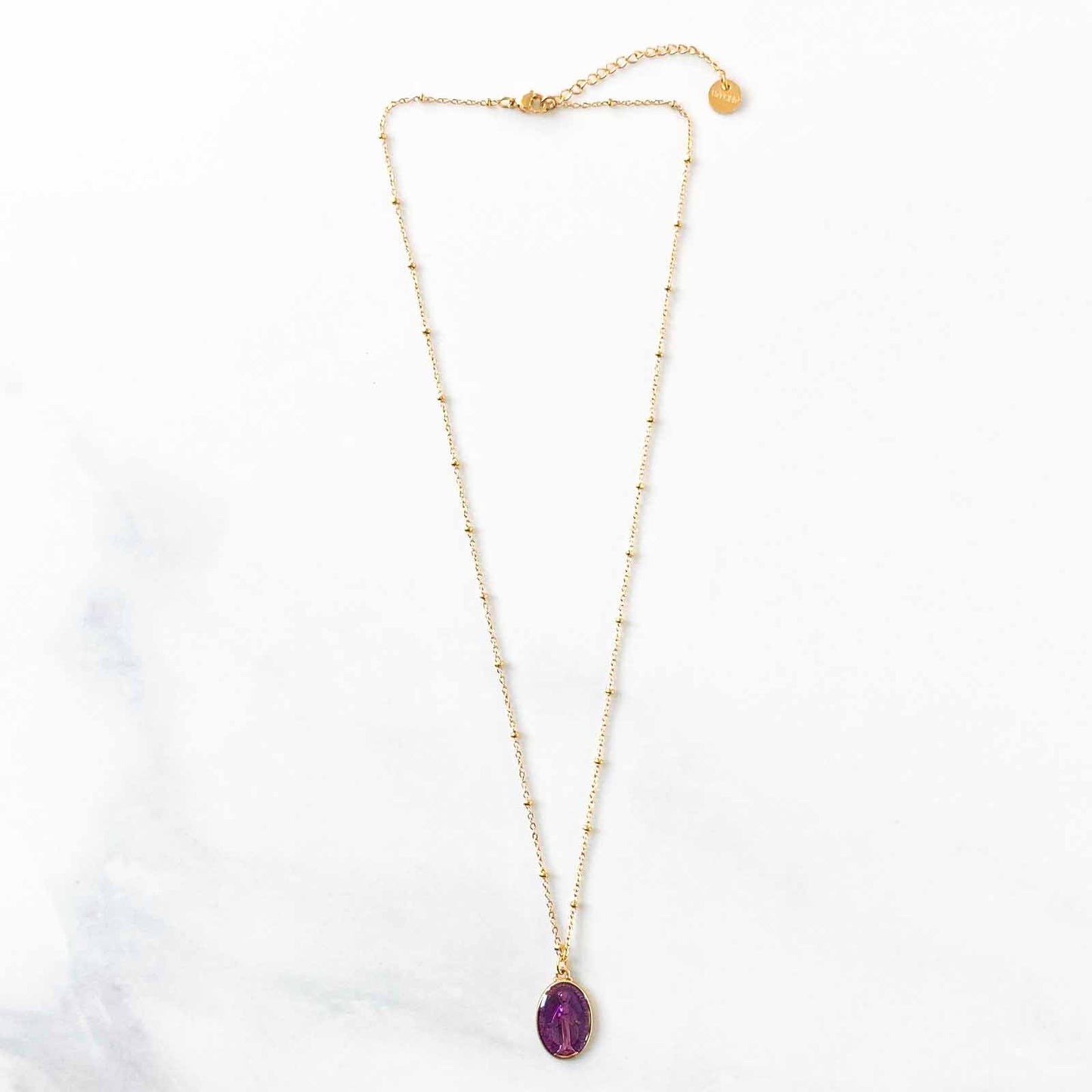 Un collier en acier inoxydable brille sous la lumière, avec des maillons lisses et robustes. Son pendentif violet, vierge de toute inscription, semble flotter délicatement, contrastant avec l'acier froid. L'ensemble incarne une élégance minimaliste, prêt à être personnalisé par celle qui le porte.