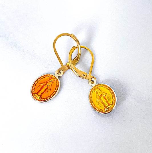 La photo capture une paire de boucles d'oreilles jaune vif, ornées d'une représentation délicate de la Vierge Marie. Les boucles d'oreilles brillent doucement sous la lumière, mettant en valeur leur couleur lumineuse. La Vierge est minutieusement détaillée, avec une expression sereine et bienveillante sur son visage. Chaque détail de ses traits est finement travaillé, ajoutant une touche de grâce à l'ensemble. Les boucles d'oreilles semblent évoquer une aura de spiritualité et de beauté divine.