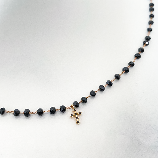 La photo représente un collier à perles noires captivant arbore un pendentif en forme de croix noir, incrusté de strass scintillants, créant un contraste saisissant. L'éclat des perles contraste avec la sobriété du pendentif, évoquant à la fois élégance et profondeur spirituelle. Ce bijou fusionne l'esthétique contemporaine avec une touche de mysticisme intemporel, captivant le regard et l'imagination.