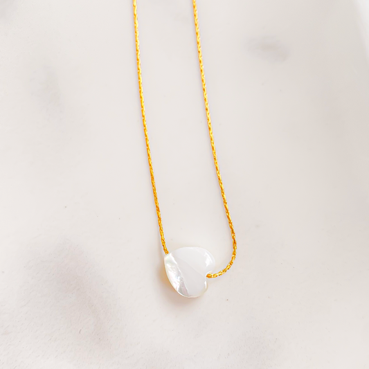 Dans la photo, un collier en forme de cœur captivant en nacre émerge avec une lueur nacrée iridescente, évoquant la douceur et l'élégance. Son design délicat et sophistiqué semble évoquer un lien intime ou un amour précieux. La lumière joue avec les nuances de la nacre, créant un effet visuel enchanteur et intemporel.