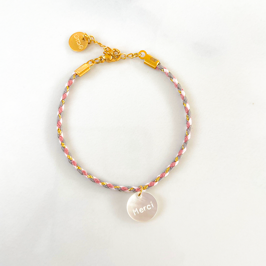 Sur la photo, un bracelet délicat en cordon parme entoure le poignet, mettant en valeur un pendentif en nacre portant le mot "merci" avec élégance. La teinte douce du parme contraste harmonieusement avec la délicatesse de la nacre, créant une pièce pleine de gratitude et de raffinement.